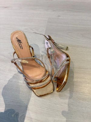 Guldiga pumps med strassdetaljer - Säljer ett par glammiga pumps från Misslucio i guld med transparenta detaljer och tunna remmar dekorerade med strass. Skorna har fyrkantig tå och justerbar rem runt ankeln. Perfekta för dig som vill sticka ut och glänsa.