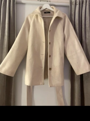 Beige kappa med bälte och knappar från Gina tricot  - Stilren beige kappa med bälte i midjan och knäppning framtill. Kappan har bred krage och raka ärmar, vilket ger en modern och clean look. Perfekt för dig som gillar minimalistisk stil och vill ha en enkel men snygg jacka till hösten eller vintern.