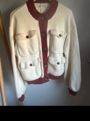 Gasta Studios Kofta  - Stilren vit kofta med vinröda ribbade muddar och knappslå. Fyra stora fickor framtill med knappar ger en cool utility-vibe. Grovstickad känsla och rund halsringning. 