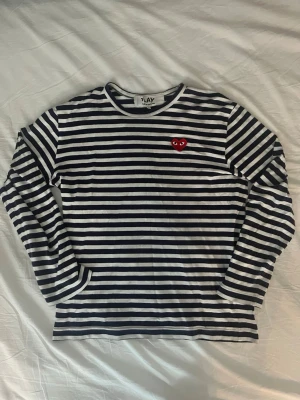 Randig långärmad tröja från Comme des Garçons - Snygg långärmad tröja med blåa och vita horisontella ränder och det klassiska röda hjärtat med ögon på bröstet. Perfekt för en stilren och trendig look. Mjuk bomullskvalitet och rund halsringning. Storlek L men sitter som m