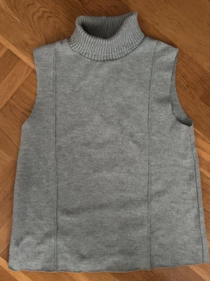 Grå stickad poloväst från Zara - Säljer en stilren grå stickad väst från Zara med hög polokrage. Västen har en rak passform och är ärmlös, perfekt att bära över en skjorta eller tröja för en trendig lager-på-lager-look.
