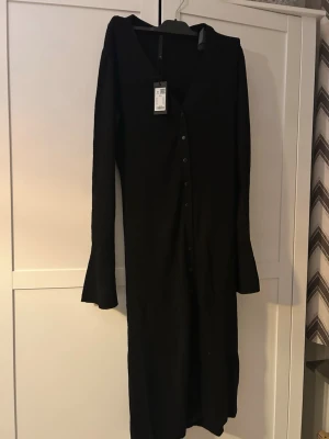 Svart ribbad långklänning med knappar - Svart långklänning med ribbad struktur och knappar längs hela framsidan. Klänningen har lång ärm och en enkel, stilren design som passar perfekt till många olika looks. Materialet ser mjukt och stretchigt ut för en bekväm passform. Helt ny!