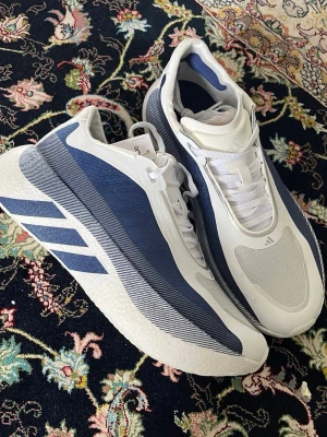 Adidas Hyperboost Edge - EU 43  - Snygga sneakers från Adidas i vitt och blått med sportig design. Skorna har lågt skaft, meshdetaljer och tydliga Adidas-ränder på sidan. Ovandelen är i textil och syntet, sulan är vit och ger en modern look. Perfekta för dig som gillar stilrena och bekväma sneakers.
