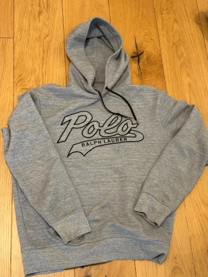 Grå hoodie från Polo Ralph Lauren - Snygg grå hoodie från Polo Ralph Lauren med stor svart logga framtill. Klassisk modell med huva och dragsko, ribbade muddar och mjukt material. Helt i nyskick 