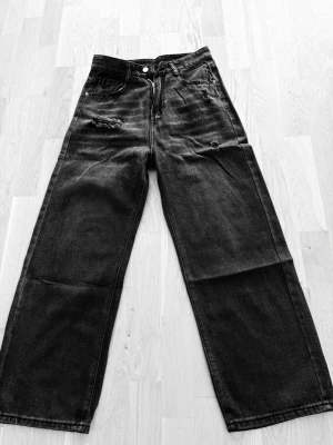 Svarta vida jeans med slitningar - Säljer ett par svarta jeans med breda ben och slitna detaljer både fram och bak. Jeansen har klassisk femficksmodell och hög midja. Materialet är kraftig denim som ger en cool och avslappnad look.