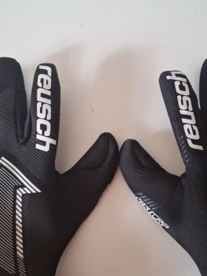 Svarta målvaktshandskar från Reusch - Säljer ett par svarta målvaktshandskar från Reusch med vit logga och detaljer. Handskarna har greppvänlig insida och är designade för att ge bra kontroll vid spel. Perfekta för fotbollsträning eller match.