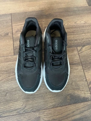 Svarta Adidas sneakers med vit sula - Säljer ett par svarta Adidas sneakers med klassiska tre ränder på sidan och vit sula. Skorna har snörning och är tillverkade i en mix av textil och syntetmaterial. Perfekta för dig som gillar en clean och sportig look.
