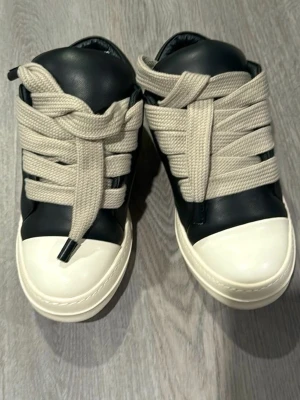 rick owens - Unika sneakers i svart och vitt med breda, krämvita snören som ger en cool och modern look. Skorna har rund tå och tjock vit sula. Materialet ser ut att vara slätt läder eller konstläder. Perfekta för dig som vill sticka ut med din stil.