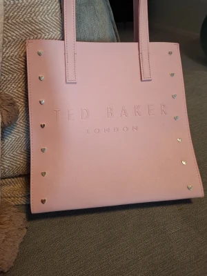 Rosa handväska från Ted Baker - Snygg ljusrosa handväska från Ted Baker med präglad logga på framsidan och små hjärtformade nitar längs kanterna. Väskan har två långa handtag och är tillverkad i ett slätt, tåligt material som ger en stilren look.