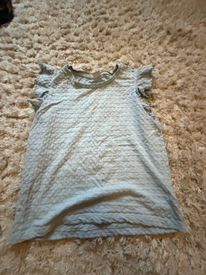 Ljusblå topp med volangärmar - Söt ljusblå topp med volangdetaljer vid ärmarna och diskret struktur i tyget. Perfekt för dig som gillar en feminin och enkel stil. Toppen är ärmlös med volang och har rund halsringning. Och de är barn storlek