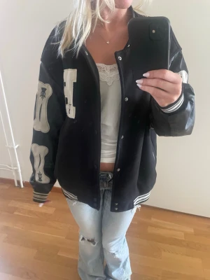 Svart bomberjacka  - Oversized svart bomberjacka med detaljer. Jackan har ribbade muddar och krage i svartvitt samt tryckknappar framtill. Ärmarna är i skinnimitation och resten i tyg. Perfekt nu till hösten! Storleken står inte i jackan men den sitter oversized på mig som är xs/s! Tveka inte att höra av er om frågor eller prisförslag!
