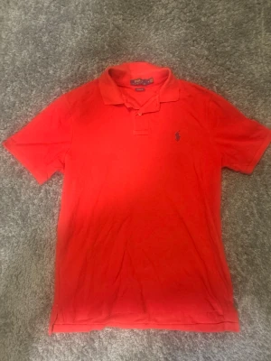 Röd pikétröja från Polo Ralph Lauren - Klassisk röd pikétröja från Polo Ralph Lauren med blå broderad logga på bröstet. Tröjan har korta ärmar, krage och knappar vid halsen. Perfekt till en avslappnad men ändå stilren look.