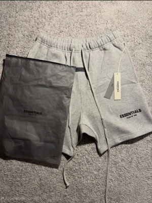 Gråa Essentials shorts med snörning - Säljer ett par gråa shorts från Fear of God Essentials med snörning i midjan och tryckt logga på benet. De har en relaxed fit och är perfekta för chill dagar. Kommer med originalpåse och tagg kvar.
