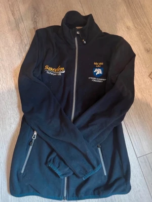 Svart fleecejacka Sweden Supporter - Svart fleecejacka med dragkedja framtill och två sidofickor med dragkedja. Broderad text 'Sweden Supporter' i gult och logga för Svenska Ridsportförbundet på bröstet. Hög krage och långärmad modell, perfekt för kyliga dagar.