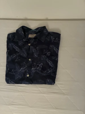 Mörkblå skjorta  från Jack and Jones - Snygg mörkblå skjorta med vita knappar och palmbladsmönster. Skjortan har klassisk krage och bröstficka, perfekt för en avslappnad och cool stil. Passar dig som gillar mönstrade plagg och vill sticka ut lite extra.
