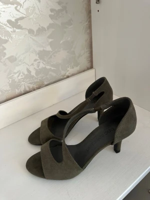 Olivgröna pumps med låg klack - Snygga olivgröna pumps från bpc selection med öppen tå och låg klack. Skorna har en elegant rem över foten och är tillverkade i mockaimitation som ger en mjuk känsla. Perfekta för dig som vill ha en stilren och bekväm sko med modern touch.