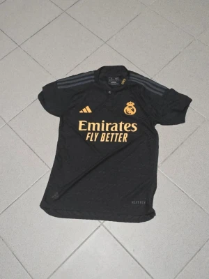 Real Madrid svart matchtröja Adidas - Svart Real Madrid matchtröja från Adidas med gula detaljer och klubbmärke. Tröjan har korta ärmar, knäppning vid halsen och tre ränder på axlarna. Perfekt för fotbollsträning eller att bära som supporter.