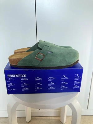 Gröna Birkenstock Boston tofflor - Snygga gröna Birkenstock Boston tofflor i mocka med justerbart spänne i metall. Klassisk korksula och mjuk fotbädd för extra komfort. Perfekta för dig som vill ha både stil och bekvämlighet i vardagen.