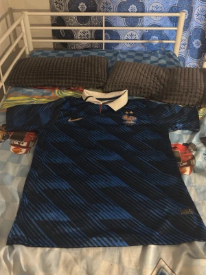 Blå och svart Frankrike fotbollströja Nike - Säljer en blå och svart fotbollströja från Nike med grafiskt mönster och vit krage. Tröjan har korta ärmar och Frankrikes landslagsmärke med två stjärnor på bröstet. Perfekt för träning eller match!