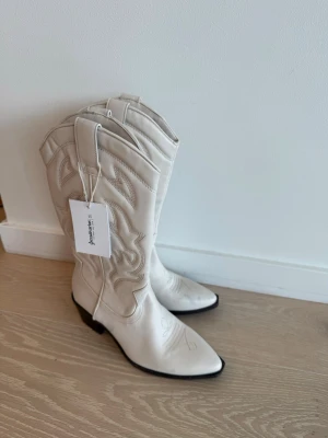 Vita cowboy boots med broderi - Snygga vita cowboy boots med klassiskt broderat mönster och spetsig tå. Bootsens skaft är medelhögt och har dragflikar upptill. Tillverkade i ett slätt material med svart sula och låg klack. Perfekta för dig som vill sticka ut med westernstil.