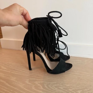 Svarta pumps med fransar och snörning Zara  - Snygga svarta pumps med höga klackar, öppen tå och coola fransdetaljer runt ankeln. Skorna har snörning som går att knyta runt benet för en trendig look. Perfekta för att lyfta vilken outfit som helst.