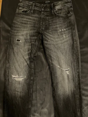 Jack and jones  - Säljer ett par svarta jeans med slitningar och distressed detaljer på framsidan. Jeansen har raka ben, fem fickor och klassisk knappgylf. Materialet är denim med en tvättad look som ger en cool och avslappnad vibe.