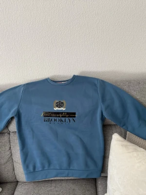 Blå sweatshirt med Brooklyn-tryck - Säljer en blå sweatshirt med tryck i svart och guld där det står 'Brooklyn', 'New York City' och 'Eighty nine'. Tröjan har rund halsringning och långa ärmar. Perfekt för en avslappnad och trendig stil.
