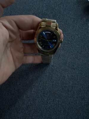 Guldig klocka med blå urtavla - Stilren klocka med guldigt metallarmband och blå urtavla. Klockan har romerska siffror och dagvisning på urtavlan. Perfekt accessoar för dig som vill sticka ut med en elegant och klassisk look.