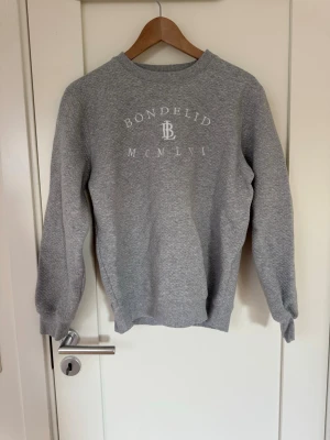 Grå sweatshirt från Bondelid - Klassisk grå sweatshirt från Bondelid med tryck framtill och rund halsringning. Tröjan har ribbade muddar vid ärmslut och nederkant samt en avslappnad passform. Perfekt för en chill och stilren look.