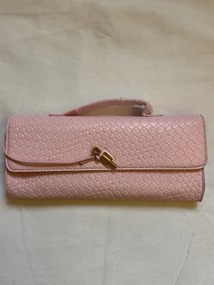 Rosa kuvertväska - Snygg rosa kuvertväska i syntetmaterial med flätad struktur och guldfärgad detalj på framsidan. Väskan har ett smalt handtag upptill och en stilren, avlång form som ger en trendig look.