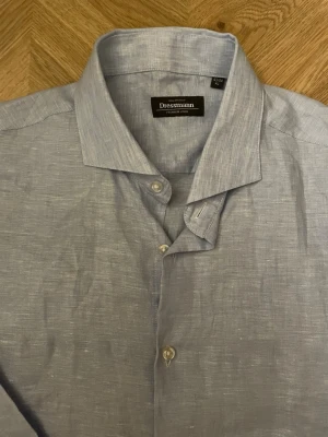 Ljusblå linneskjorta från Dressmann - Snygg ljusblå skjorta från Dressmann i luftigt linne. Klassisk krage, knappar framtill och långa ärmar med manschett. Perfekt för en stilren och avslappnad look.