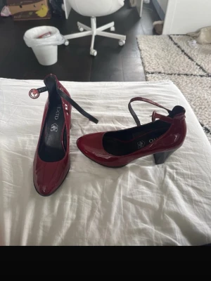 Vinröda lackpumps med rem - Säljer ett par vinröda äkta skinn klackskor  från Texto i glansigt lackmaterial. Skorna har en rundad tå, bred klack och en justerbar rem runt ankeln. Snygg kontrast med svart sula och klack. Perfekta för dig som vill sticka ut med färg.