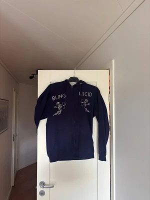 Mörkblå Zip från Bling Lucid - Mörkblå zip från Bling Lucid. På bröstet står det 'BLING LUCID' med rhinestones tryck av två änglar. Hoodien har fickor framtill och en avslappnad passform, perfekt för en chill stil.