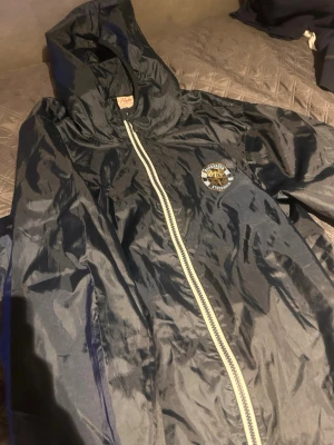 Svart vindjacka med Djurgården-tryck - Snygg mörkblå vindjacka med huva och vit dragkedja. På bröstet finns ett tryck med Djurgården Stockholm-emblem i guld, ljusblå och blått. Jackan är tillverkad i ett glansigt, tunt material som passar perfekt för blåsiga dagar.