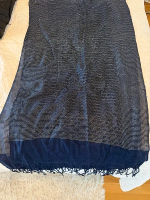 Mörkblå sjal med tunna ränder - En stor, tunn mörkblå sjal med tunna horisontella ränder i guldfärg.. Sjalen har fransar längs kanterna och är tillverkad i ett lätt och luftigt material som känns skönt mot huden. Perfekt att slänga över axlarna för en snygg touch.