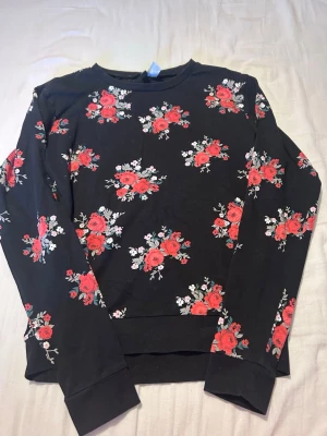 Svart sweatshirt med röda blommor - Svart långärmad sweatshirt med tryck av röda rosor och små vita samt rosa blommor över hela tröjan. Rund halsringning och ribbade muddar vid ärmslut och nederkant. Perfekt för dig som gillar blommiga mönster och vill sticka ut lite extra.