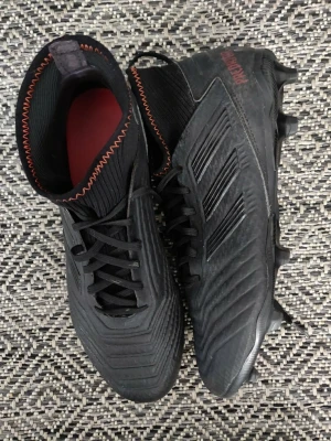 Svarta fotbollsskor med högt skaft - Säljer ett par helsvarta fotbollsskor från Adidas predator med högt skaft och orangea sömmar. Skorna har snörning, mönstrad ovandel och fasta dobbar för spel på gräs.  Kan även hämtas i Stockholms stad.