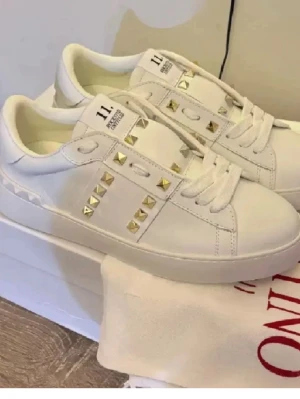 Vita sneakers med gulddetaljer - Stilrena vita sneakers i läder med guldfärgade nitar längs sidorna och på hälen. Skorna har tjock vit sula med rutmönster undertill och vita skosnören. Märket syns på plösen och designen är modern och edgy.