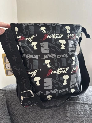 Snoopy axelväska med tryck - Cool svart axelväska med justerbar rem och tryck av Snoopy, texten 'Joe Cool' och olika grafiska detaljer i vitt, grått och rött. Väskan har en rektangulär form och är tillverkad i slitstarkt tygmaterial. Perfekt för dig som gillar unika accessoarer med karaktär. Mått 32x28x10 cm 