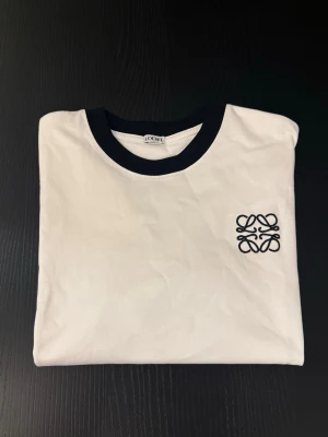 Vit t-shirt med svart logga från Loewe oanvänd - Vit t-shirt från Loewe med svart ribbad krage och svart broderad logga på bröstet. Klassisk passform och korta ärmar. Perfekt till jeans eller shorts för en clean och stilren look.