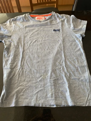 Grå t-shirt från Superdry - Snygg grå t-shirt från Superdry med rund halsringning och diskret Superdry-logga i blått på bröstet. Tillverkad i mjuk bomull för en skön känsla. Perfekt till jeans eller shorts för en avslappnad look. Vintage snygg.