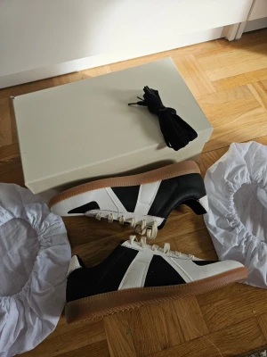 Maison Margiela Oreo Gats - Snygga Maison Margiela Oreo Gat sneakers i svart och vitt tyg med klassiska vita detaljer och snörning. Skorna har en brun gummisula som ger en retrovibe och kommer med extra svarta skosnören. Perfekta för dig som gillar stilrena och sportiga sneakers.