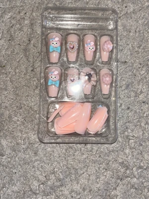 Söta rosa press-on naglar med dekorationer - Supergulliga press-on naglar i ljusrosa med 3D-dekorationer som nallebjörnar, rosetter, pärlor och hjärtan. Naglarna har en blank finish och varierande dekorationer i blått och rosa. Perfekt för dig som vill ha en söt och unik nagelstil.