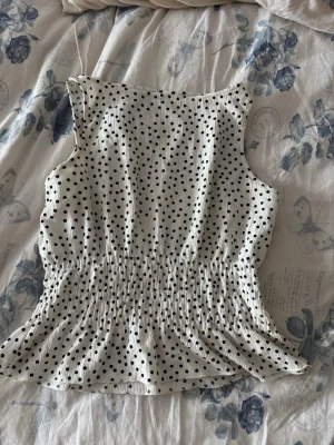 Vit prickig ärmlös topp - Säljer en söt vit topp med svarta prickar. Toppen är ärmlös och har en smockad midja som ger en snygg passform och volangkant nedtill. Perfekt för dig som gillar en lekfull och trendig stil.