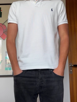 Vit pikétröja från Polo Ralph Lauren - Klassisk vit pikétröja från Polo Ralph Lauren i slim fit-modell. Tröjan har korta ärmar, krage och knappar vid halsen samt den ikoniska broderade loggan på bröstet i marinblå färg.