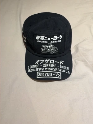 Svart keps med japansk text och brodyr - Säljer en svart supreme keps med broderade vita texter på japanska och engelska samt en 4-hjuling på framsidan. Kepsen har böjd skärm och detaljer på både fram- och sidopanelen. Perfekt för dig som gillar streetwear och unika accessoarer.