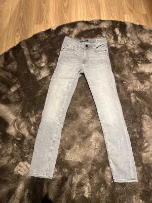 Ljusgråa slim jeans - Säljer ett par ljusgråa slim jeans med klassisk femficksdesign och smal passform. Jeansen har dragkedjegylf och bälteshällor. Snygga detaljer på bakfickorna och passar perfekt till en avslappnad stil. 27/30