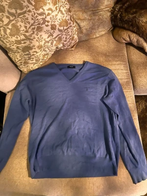Blå V-ringad tröja från Gant - Snygg blå V-ringad tröja från Gant i tunnare material. Tröjan har långa ärmar, ribbade muddar och en diskret broderad logga på bröstet. Perfekt att bära över en skjorta eller t-shirt för en clean och stilren look.