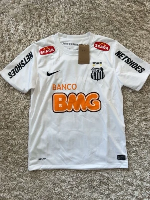 Vit Santos Neymar Jr matchtröja Nike - Säljer en vit Santos matchtröja från Nike med Neymar Jr och nummer 11 på ryggen. Tröjan har flera sponsorloggor i svart, rött och orange samt klubbmärke på bröstet. Tillverkad i lätt och ventilerande material, perfekt för fotbollsträning eller match.