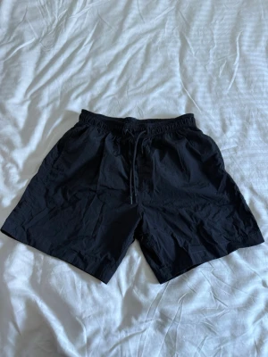 Svarta badbyxor från H&M - Säljer ett par klassiska svarta badbyxor från H&M i storlek S. De har elastisk midja med dragsko och meshfoder på insidan för extra komfort. Har även innerbyxa i mesh och en bakficka med kardborrestängning. Perfekta för stranden eller poolen.